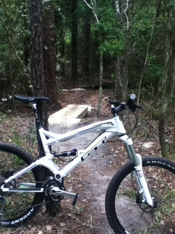 A mountain bike parked beside a dirt trail in a wooded area, with trees and underbrush surrounding it. A wooden ramp is visible in the background, suggesting a biking trail designed for challenging terrain. Tillie Fowler Regional Park mountain bike trail.