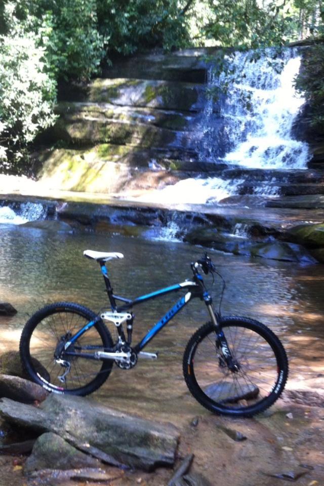 Trek Fuel EX 9.8: A mountain bike resting on rocky ground near a clear stream, with a waterfall cascading in the background, surrounded by lush green foliage.