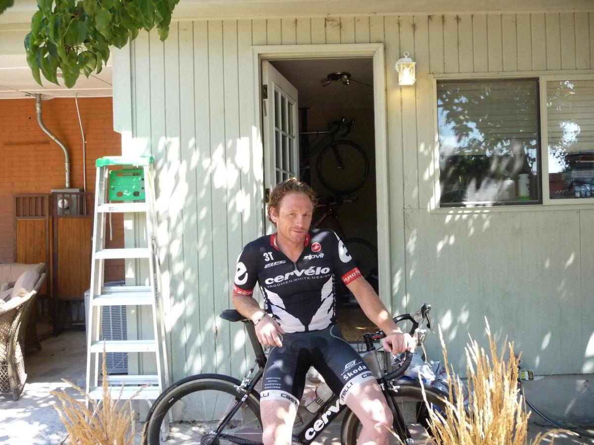 Cannondale Scalpel 1: A cyclist in a black and white cycling jersey sits on a bike outside a green house with a sunny patio. Behind him, a ladder and a few bicycles can be seen. The setting has a relaxed, outdoor atmosphere with some plants in the foreground.