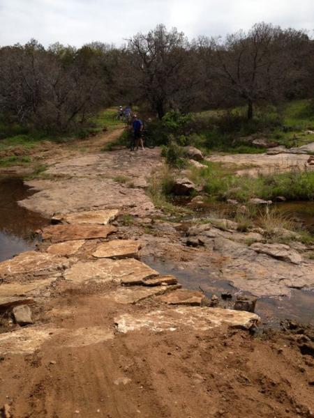 A rocky path crosses a small stream in a natural outdoor setting, surrounded by sparse trees and greenery. Two people can be seen in the background, one of whom appears to be adjusting a bicycle. The ground is a mix of dirt and stones, suggesting a scenic hiking or biking trail. Reveille Peak Ranch mountain bike trail.