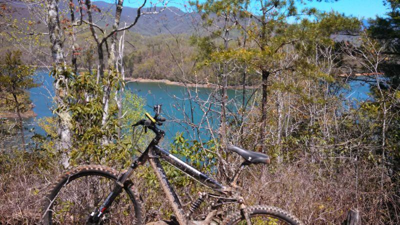 A mountain bike leaning against a tree, with a scenic view of a turquoise lake and mountains in the background, surrounded by green foliage and trees. Tsali Mouse Branch Loop mountain bike trail.