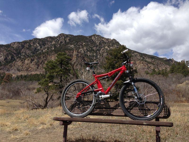A bright red mountain bike positioned on a metal bench, with a scenic backdrop of rugged mountains and blue skies dotted with white clouds. Surrounding the bench are patches of dry grass and sparse trees, illustrating a serene outdoor setting. Cheyenne Mountain State Park mountain bike trail.