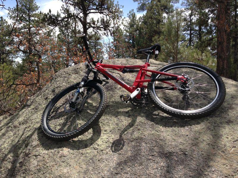 A bright red mountain bike rests on a large rock in a forested area, surrounded by tall trees and a partly cloudy sky. The bike is positioned at an angle, showcasing its wheels and frame against the natural backdrop. Cheyenne Mountain State Park mountain bike trail.