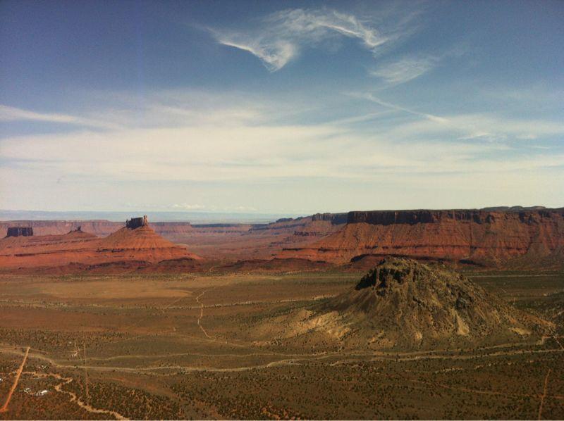 A panoramic view of a desert landscape featuring red rock formations and mesas under a bright sky with scattered clouds. In the foreground, a small hill is visible, while the vast valley stretches out with a mix of vegetation and dirt paths leading into the distance. Porcupine Rim mountain bike trail.