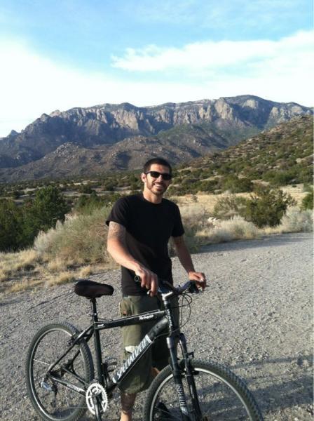 A man standing next to a black mountain bike on a gravel path, with a mountainous landscape in the background and a clear blue sky. He is smiling and wearing sunglasses, dressed in a black t-shirt and green cargo shorts. Sparse vegetation and hills surround him. Sandia Mountains Foothill Trail mountain bike trail.
