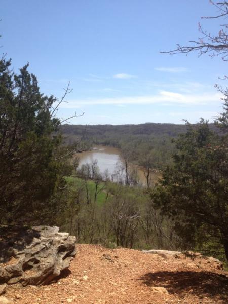 A scenic view from a rocky overlook, showcasing a winding river surrounded by trees and greenery under a clear blue sky. Castlewood State Park mountain bike trail.