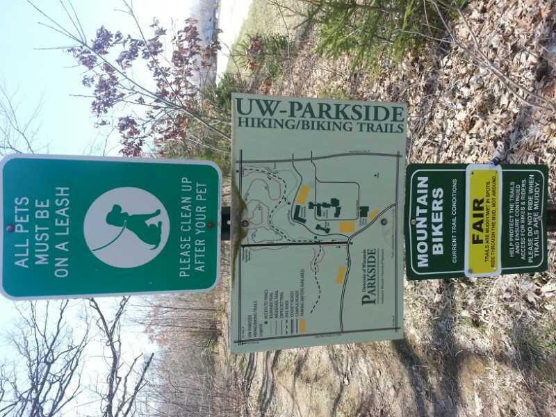 Signage at UW-Parkside indicating hiking and biking trails. The left sign instructs that all pets must be on a leash and encourages owners to clean up after their pets. The center sign features a map of the trails, while the right sign provides information for mountain bikers regarding current trail conditions, indicating that they are rated as "Fair." Surrounding area includes trees and natural terrain. Petrifying Springs mountain bike trail.