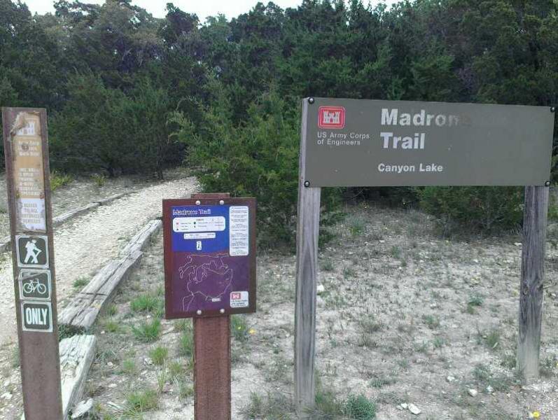Signage at the entrance of the Madrone Trail near Canyon Lake, featuring information about trail use for hikers and cyclists. The path is visible leading into a wooded area, with trees in the background. Madrone Trail mountain bike trail.