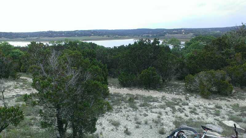 A scenic view of a landscape featuring a lake in the distance, surrounded by lush greenery and scattered trees. The foreground includes rocky terrain with patches of grass, while the sky is overcast, suggesting a calm and cool atmosphere. A bicycle is partially visible on the ground, indicating an outdoor recreational activity. Madrone Trail mountain bike trail.