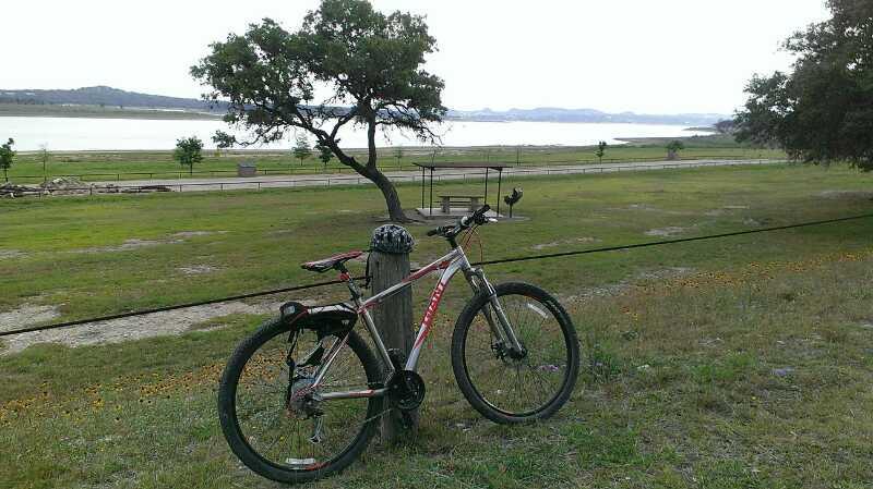 A mountain bike leaning against a wooden post in a grassy area, with a lake and distant hills visible in the background. The scene is peaceful and showcases nature, with a tree providing shade. Madrone Trail mountain bike trail.