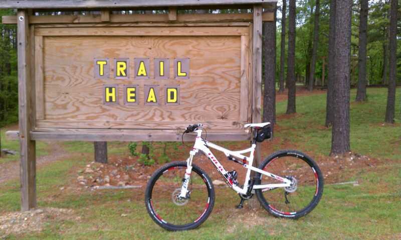 A white mountain bike parked next to a wooden sign that reads "TRAIL HEAD" in yellow letters, set against a backdrop of trees and grassy terrain. Cedar Glades Trail mountain bike trail.