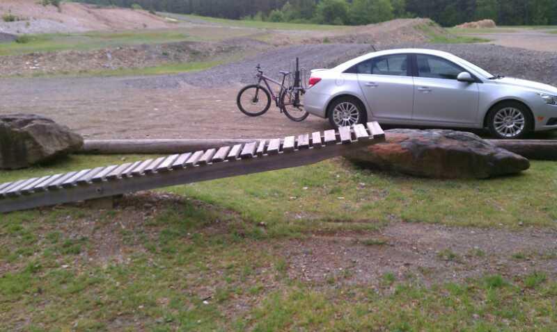 A wooden bike ramp extends over a rocky area, leading from the ground to a large boulder. In the background, a silver car is parked nearby, with a bicycle mounted on a rack attached to the back of the vehicle. The setting appears to be an outdoor area with gravel and grass, surrounded by trees. Cedar Glades Trail mountain bike trail.