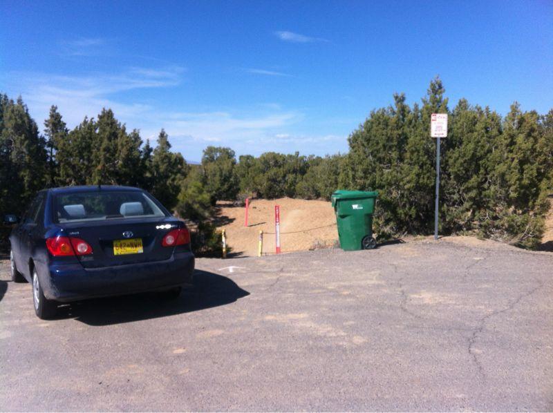 A blue car parked on a gravel lot with green bushes and trees in the background. A green trash bin is nearby, and there is a sign indicating no parking. The sky is clear with a few clouds. The Whoops mountain bike trail.