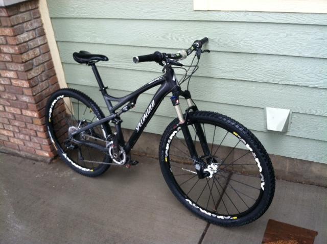 Specialized Epic Comp 29er: A black mountain bike leaning against a textured green wall, with a brick column in the background. The bike features thick tires, a front suspension fork, and a modern frame design, suggesting it is suitable for rough terrain. The ground is wet, indicating recent rain.