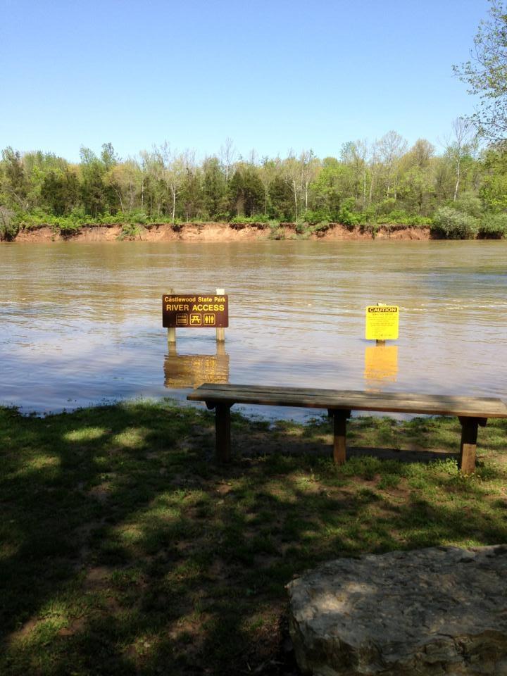 A view of a river access area at Castlewood State Park on a clear day. Two signs are visible near the water: one indicating "RIVER ACCESS" with symbols for fishing and restrooms, and another caution sign. The river is calm, with trees lining the opposite bank and a grassy area in the foreground. A bench is positioned near the water's edge. Castlewood State Park mountain bike trail.