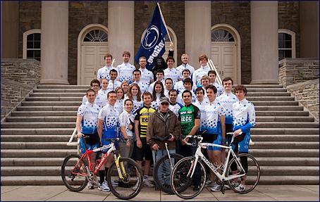 A group photo of a cycling team gathered in front of a historic building. The team members are wearing matching white jerseys with blue accents, and they are positioned around a central figure holding a bicycle. There are two bicycles displayed prominently in the foreground. A larger team flag is held at the back, adding to the team spirit.