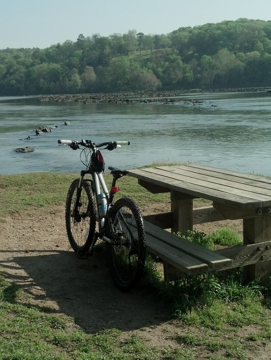 A mountain bike parked next to a wooden picnic table, with a scenic view of a river and lush green trees in the background. The peaceful outdoor setting is bathed in natural sunlight. Augusta Canal mountain bike trail.