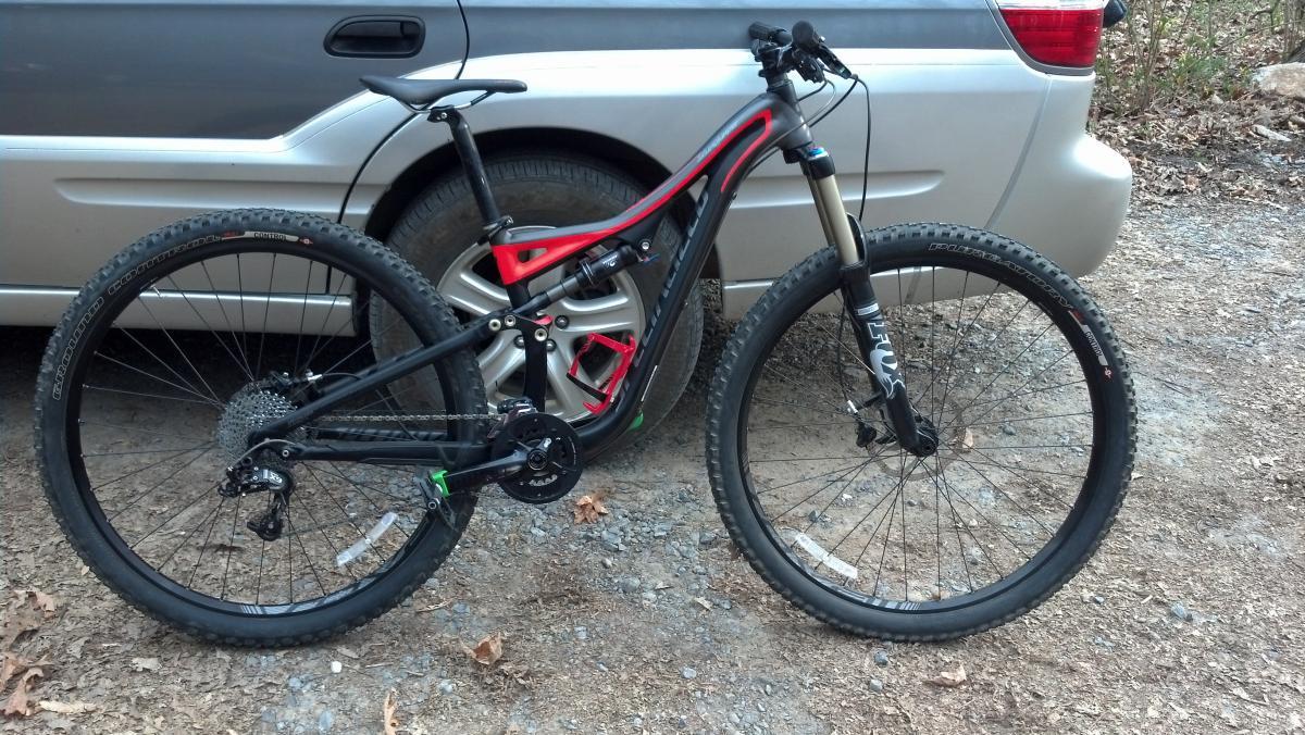 Specialized Stumpjumper FSR Comp 29: A black and red mountain bike leaning against a silver car, parked on a gravel surface surrounded by autumn leaves. The bike features thick tires designed for off-road riding and a modern frame with noticeable suspension components.