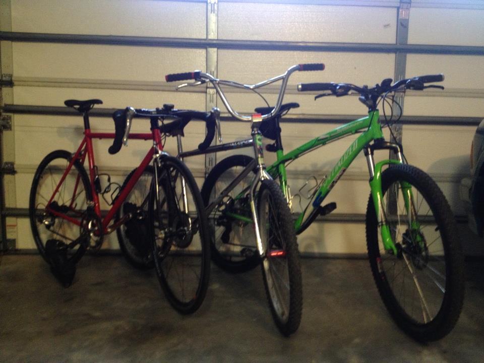Specialized Hardrock Disc: Two bicycles, one red road bike and one green mountain bike, are parked next to each other in a garage with a closed garage door in the background. The red bike features drop handlebars and thin tires, while the green bike has wider tires and a more upright handlebar design.