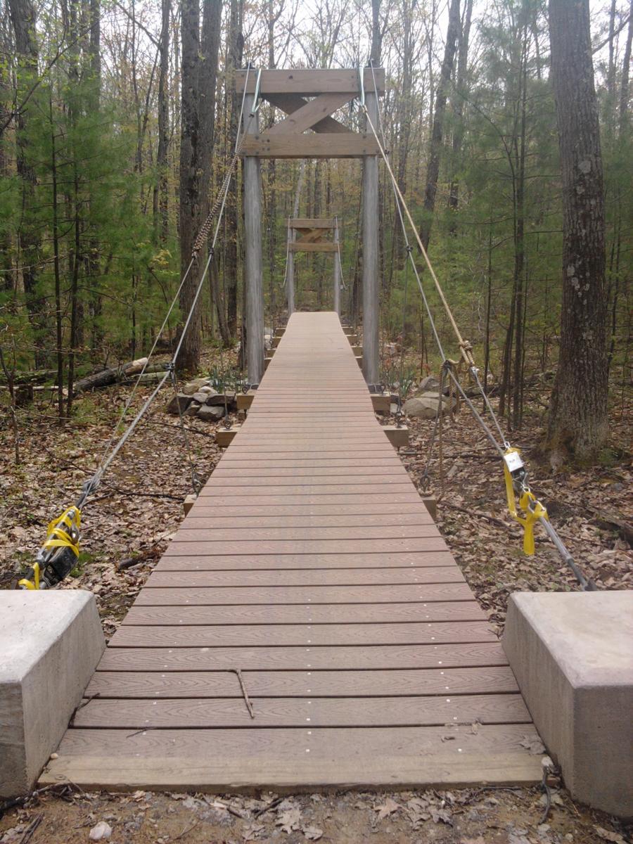 A wooden suspension bridge stretches through a forest, with sturdy wooden supports and cables. The path is surrounded by trees and scattered leaves, leading into a serene natural setting. Pine Hill Park mountain bike trail.