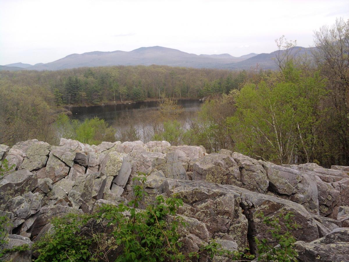 A rocky outcrop in the foreground overlooking a calm lake surrounded by lush greenery and rolling hills in the background, under a cloudy sky. Pine Hill Park mountain bike trail.