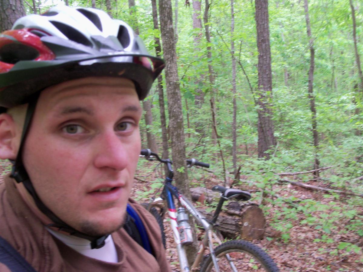 A person wearing a bike helmet is standing in a lush forest, looking towards the camera with a concerned expression. A mountain bike is positioned nearby among the trees and foliage, creating an outdoor biking scenario. Lynches Woods mountain bike trail.
