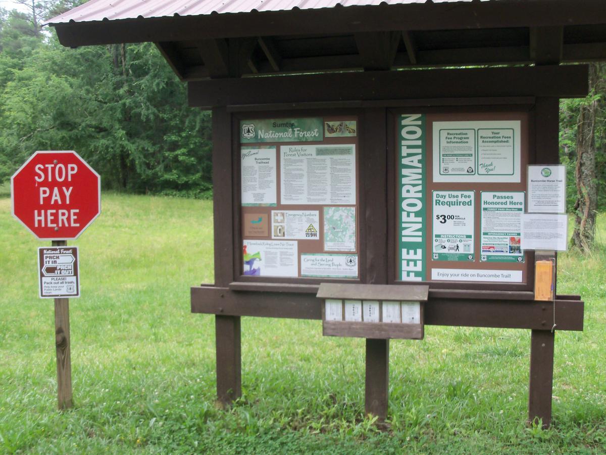A wooden information board at a national forest featuring notices about recreational fees, including a "Day Use Fee Required" sign. Nearby, a red stop sign instructs visitors to "STOP PAY HERE." The board includes various informational posters, maps, and instructions, set against a backdrop of greenery. Buncombe - Brickhouse Recreation Area mountain bike trail.