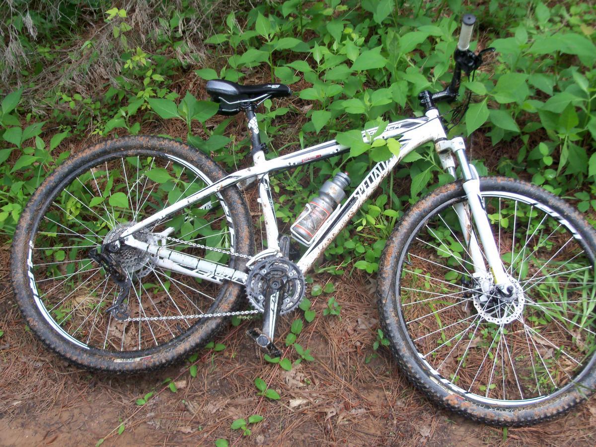 A white mountain bike lying on the ground, partially covered in dirt, amidst green foliage and pine needles. A water bottle is mounted in the frame, and the bike appears to have recently been used for trail riding. Buncombe - Brickhouse Recreation Area mountain bike trail.