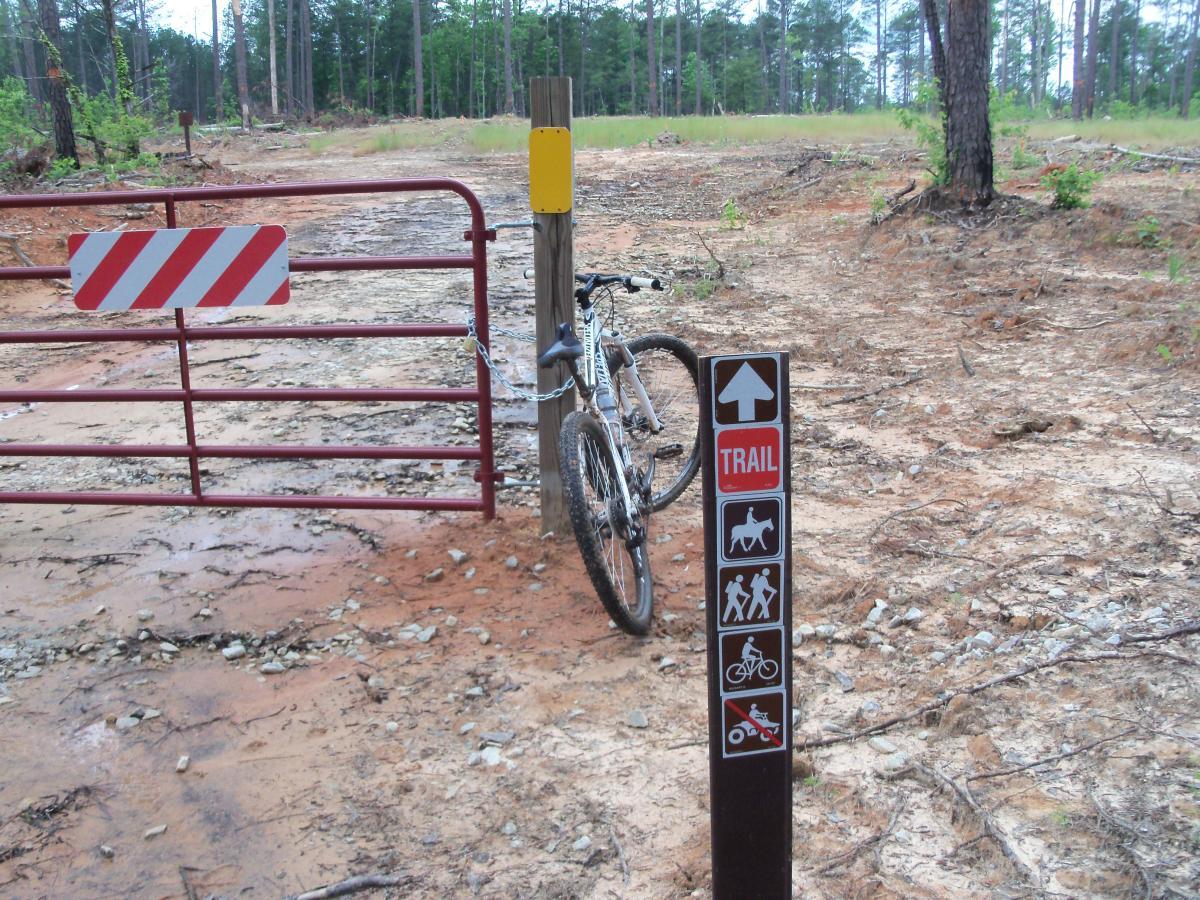 A mountain bike is leaned against a wooden post near a closed metal gate with red and white stripes. In the foreground, there is a trail marker sign indicating permitted activities, including hiking, biking, and horseback riding. The background features a dirt path leading through a forested area with sparse vegetation and pine trees. Buncombe - Brickhouse Recreation Area mountain bike trail.