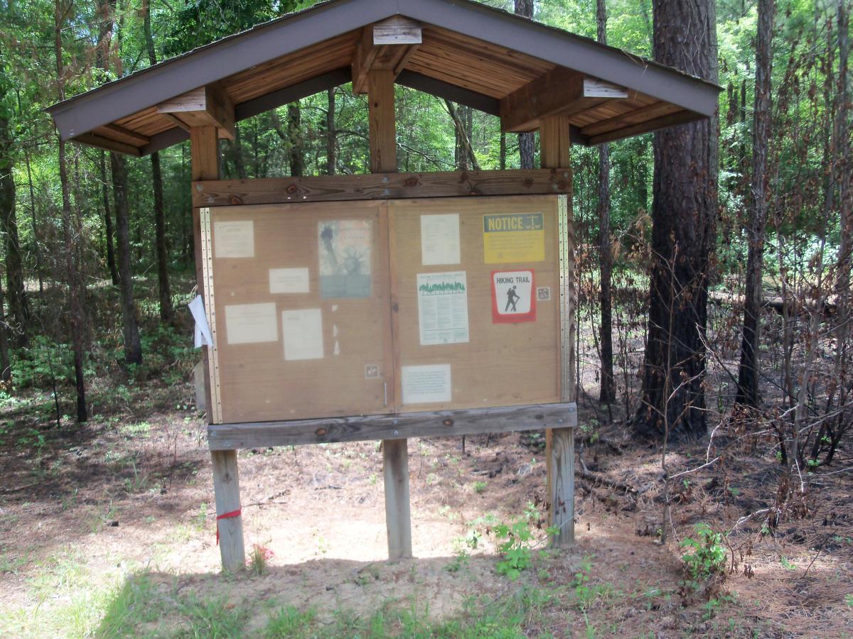 A wooden information board with a roof, displaying various notices and signs, situated in a wooded area. The board includes a "Hiking Trail" sign, a warning notice, and multiple informational sheets. Surrounding vegetation shows signs of recent burn, with some charred plants visible in the background. Wine Creek mountain bike trail.