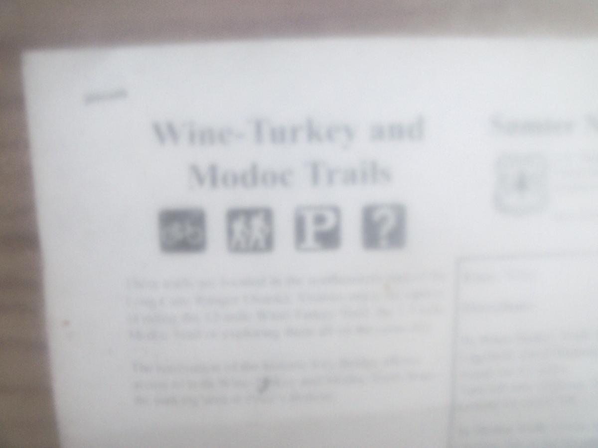 Image of a informational sign titled "Wine-Turkey and Modoc Trails." The sign features icons representing various activities, including hiking and parking. The background contains text that provides information about the trails and nearby facilities. The image is slightly blurred, making some details less clear. Wine Creek mountain bike trail.