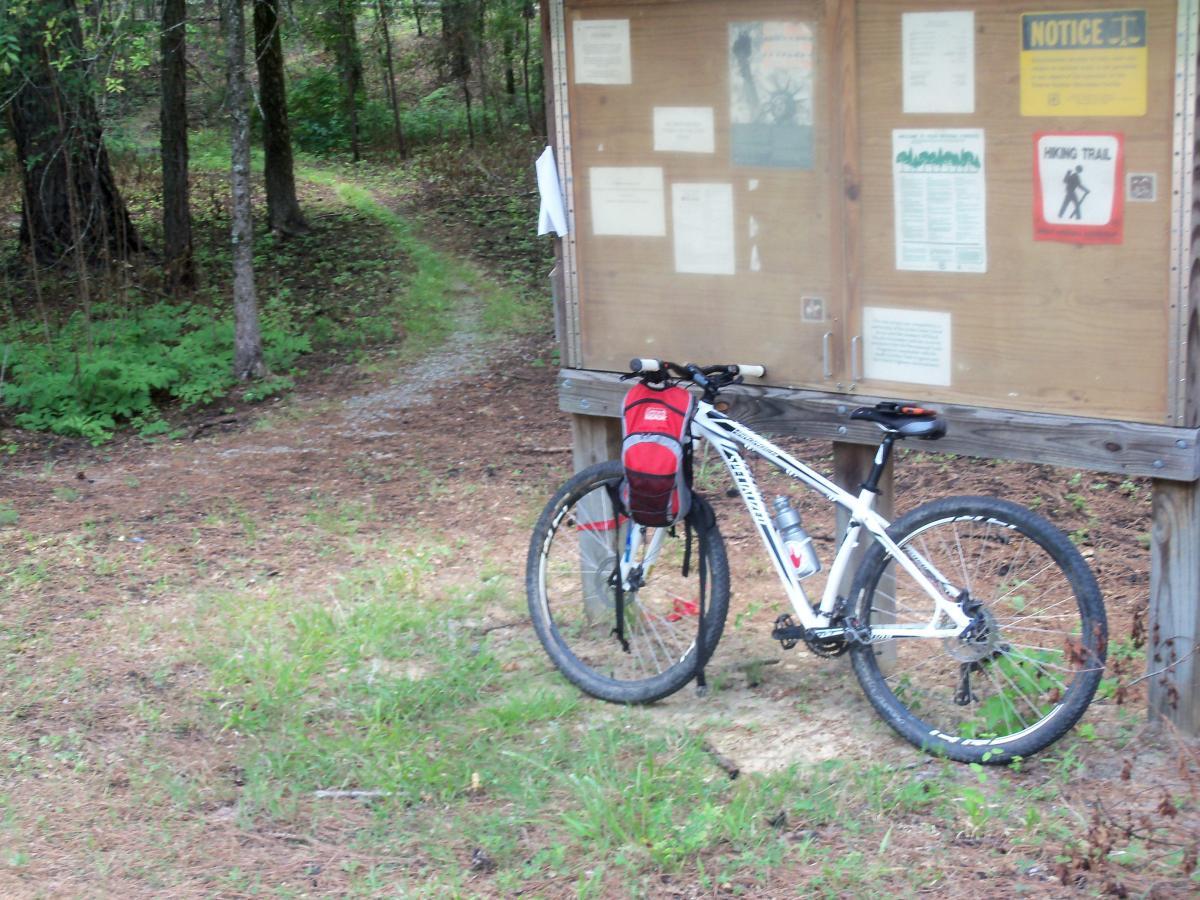 A mountain bike leaning against a wooden information board in a forested area, with a gravel path visible leading into the woods. The board displays various signs and notices about the trail. Surrounding vegetation includes trees and patches of grass. Wine Creek mountain bike trail.