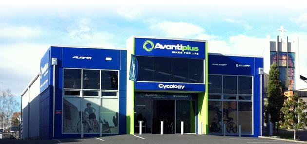 Exterior view of an Avantiplus bike shop, featuring blue and green signage that reads "Avantiplus Bikes for Life" and "Cyclology." The building has large windows showcasing bike displays and a modern architectural design, situated in a well-maintained area. Clear skies and a few clouds can be seen in the background.