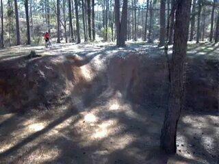 A dirt bike rider in a red shirt stands near a steep, sandy embankment surrounded by tall trees in a forested area. Sunlight filters through the branches, casting shadows on the ground. Flat Rock Park mountain bike trail.