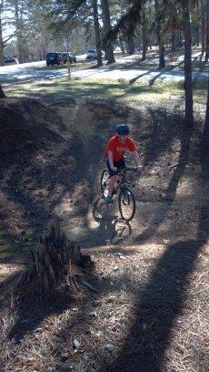 A person riding a mountain bike on a dirt trail surrounded by trees, with sunlight filtering through the branches. The rider is wearing a blue helmet and an orange t-shirt, navigating a small incline near a tree stump. Flat Rock Park mountain bike trail.