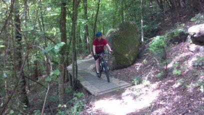 A person riding a mountain bike on a wooden bridge surrounded by dense green trees and large rocks in a forested area. Chewacla State Park mountain bike trail.