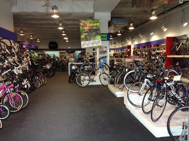 Interior of a bike shop showcasing a wide variety of bicycles. The store is well-lit, with bikes displayed along the walls and in the center aisle. Shelves contain biking accessories and products. A sign promoting fitness and biking hangs from the ceiling.