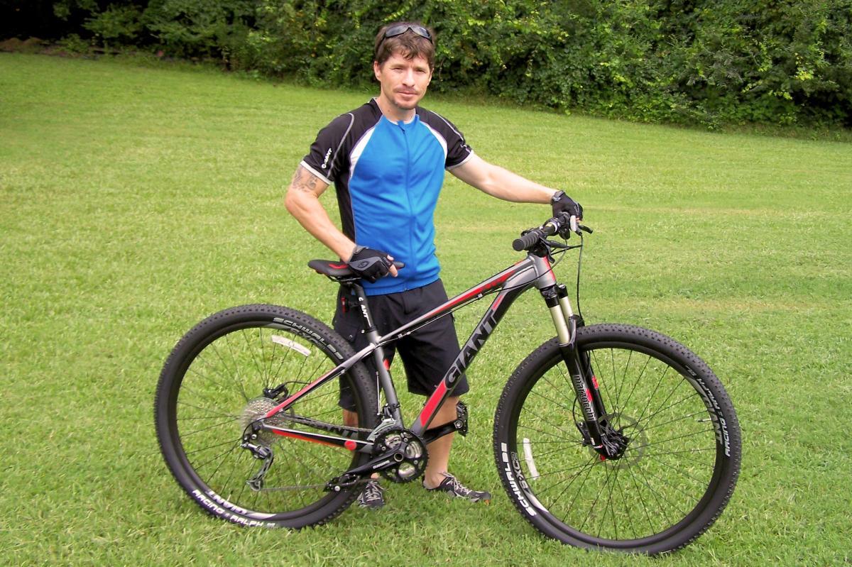 Giant XTC 29er 2: A person standing on grass beside a mountain bike, wearing a blue and black cycling jersey and black shorts. The bike has a prominent "Giant" logo and features thick tires, suitable for off-road riding. The background is lush greenery, suggesting an outdoor setting.