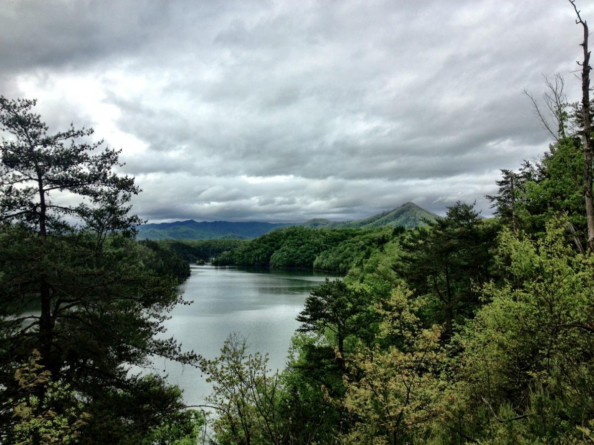 A tranquil view of a lake surrounded by lush green trees and rolling hills under a cloudy sky. The water reflects the scenery, with distant mountains visible in the background, creating a serene natural landscape. Tsali Recreation Area mountain bike trail.