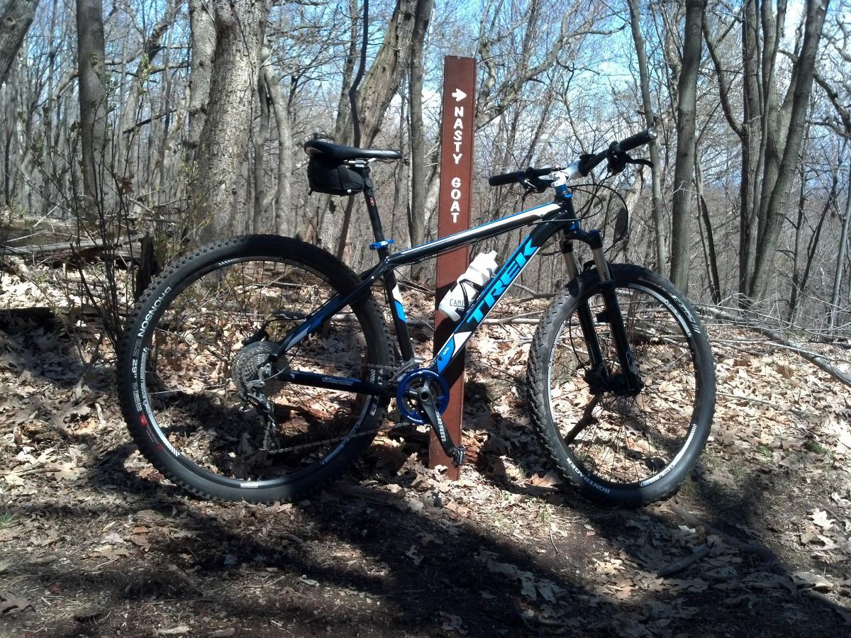 A mountain bike leaning against a trail sign labeled "Nasty Goat," surrounded by trees and fallen leaves. The bike features a blue and black frame, with sturdy tires suitable for rugged terrain. The background shows a wooded area with bare branches, suggesting early spring or late fall. Northwest Park mountain bike trail.