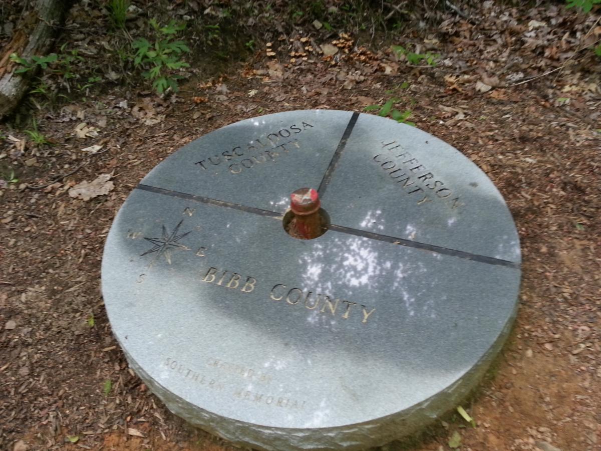 A circular stone marker on the ground, indicating the intersection of four counties: Tuscaloosa County, Bibb County, Jefferson County, and a fourth unnamed area. The marker has a compass rose in the center and is surrounded by a natural forest floor with leaves and small plants. Tannehill Historic Ironworks State Park mountain bike trail.