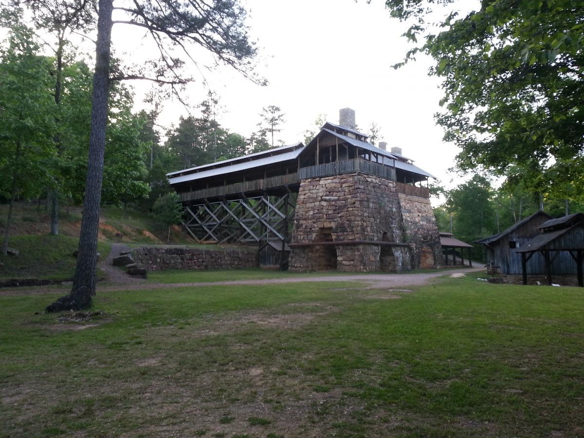 A large, rustic stone building elevated on wooden stilts, surrounded by lush green trees and grass. A winding dirt path leads towards the structure, which features a sloped roof and a balcony on the upper level. To the right, several smaller wooden buildings are visible, adding to the serene natural setting. Tannehill Historic Ironworks State Park mountain bike trail.