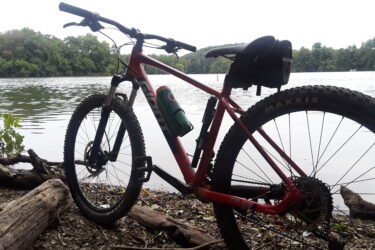 A mountain bike resting on the shore near a calm lake, surrounded by trees and natural scenery. The bike, with a red frame and black tires, is positioned to show the front wheel and handlebars. A water bottle is attached to the frame, and there are some logs and stones in the foreground. The sky is overcast, suggesting a serene outdoor atmosphere. Blue Marsh mountain bike trail.