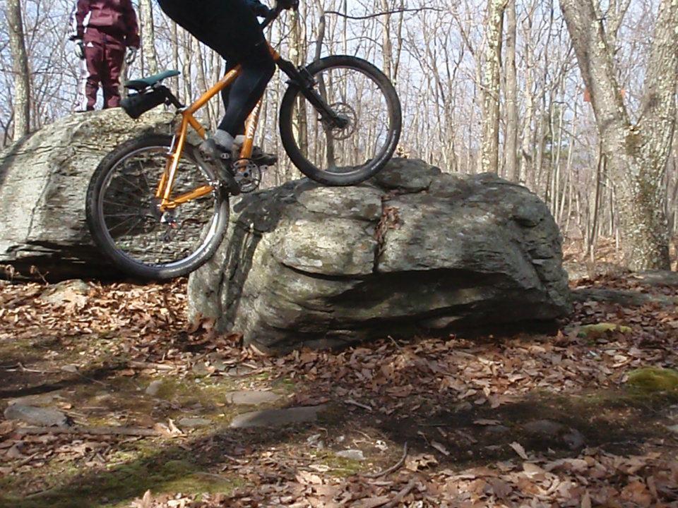 A mountain biker negotiating a large rock while riding through a wooded area, with fallen leaves scattered on the ground. Another person is standing in the background, observing the biking maneuver. Moon Lake Park mountain bike trail.
