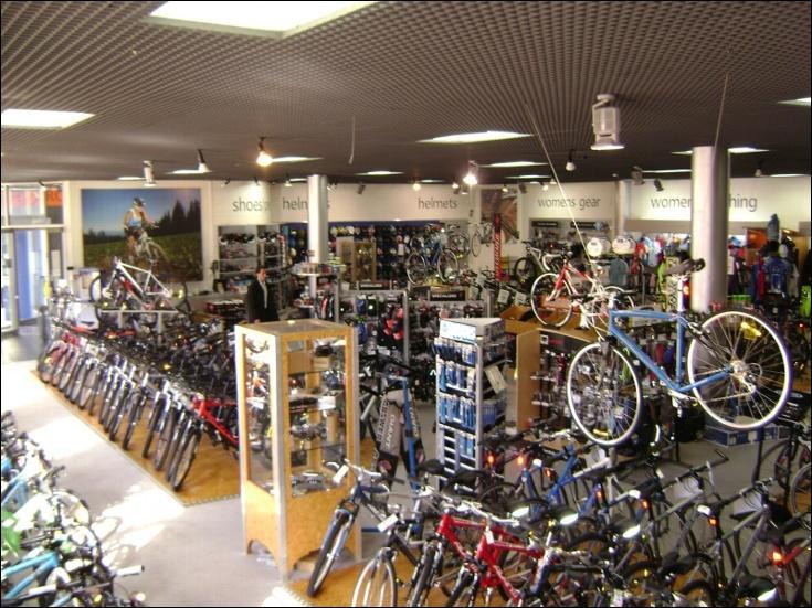 Alt text: A spacious bicycle shop filled with various models of bikes displayed on the floor and hanging from the ceiling. The store features sections for helmets, shoes, women