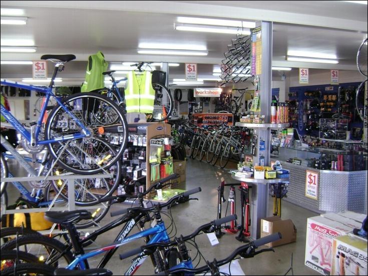 A bicycle shop interior filled with various bicycles, bike accessories, and tools. The space features a selection of colorful bikes displayed prominently, shelves stocked with gear, and clearly marked price tags. Bright lighting enhances the organized layout, with a workshop area visible in the background.