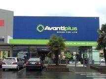 Front view of an Avantiplus store featuring a large blue sign with their logo and slogan "Bikes for Life." The building has glass doors and windows, with vehicles parked in front, and a palm tree visible near the entrance. The sky appears overcast.