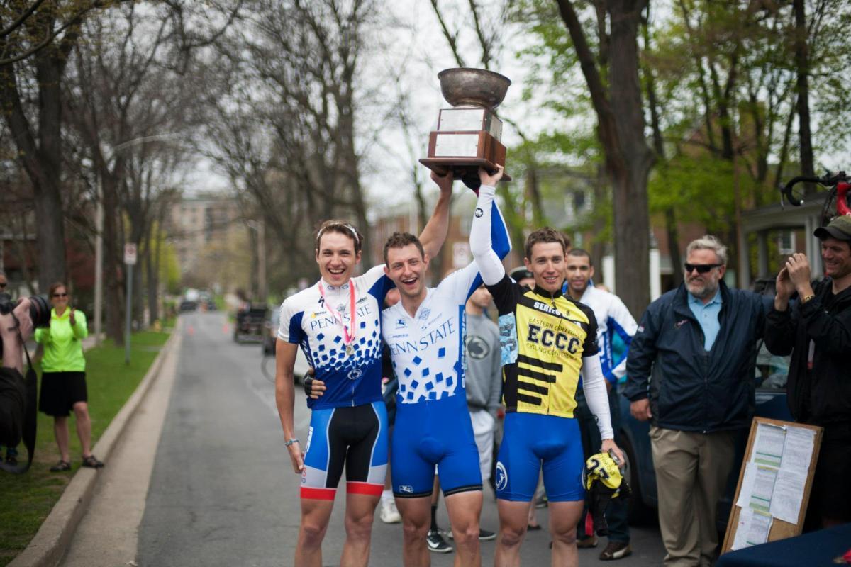 Three cyclists celebrate their victory, with one holding a large trophy above their heads. They are dressed in cycling jerseys, with two wearing blue and white gear representing Penn State and one in a yellow and black jersey from the Eastern Collegiate Cycling Conference. Surrounding them are spectators and fellow cyclists, with trees lining the street in the background. The atmosphere is joyous and festive, marking a significant achievement in a cycling event.