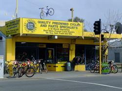 A bright yellow bicycle shop with a sign that reads "Quality Preowned Cycles and Parts Specialists." The storefront features several bicycles outside, and one bicycle is hanging from the roof. There is a traffic light and street signs visible in the foreground.
