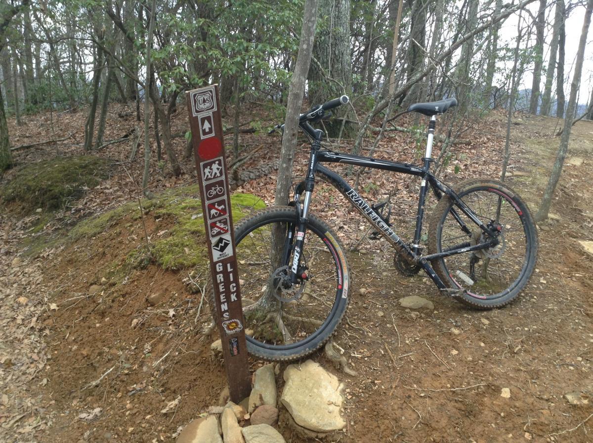 Raleigh Mojave 8.0: A black mountain bike leans against a trail sign in a wooded area, with dirt and rocks on the ground. The sign features several symbols indicating recreational activities, including hiking and biking, and displays the name "Greens Licks." The trees in the background are bare, suggesting a spring or fall season.